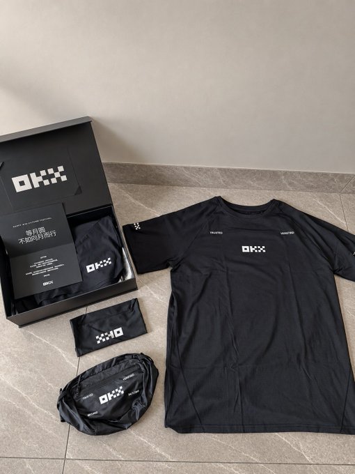 A black OKX branded t-shirt is displayed alongside its packaging box and accessories on a tiled floor.