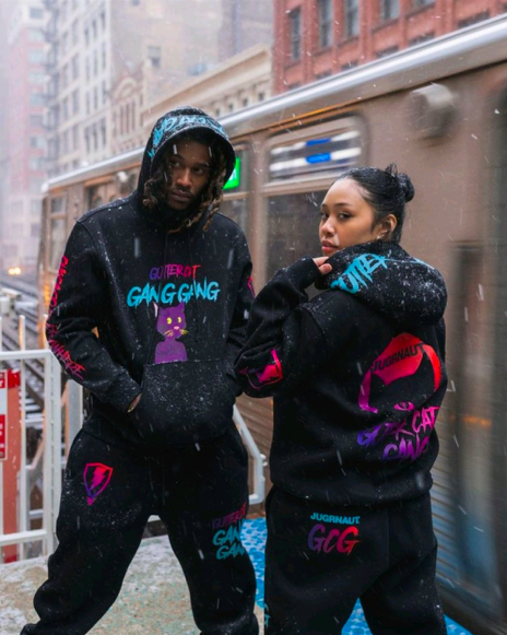 Two people in a snowy city street wearing matching black hoodies and sweatpants with colorful graffiti-style graphics including 'GUERROT GANG GANG' and 'G2G'.