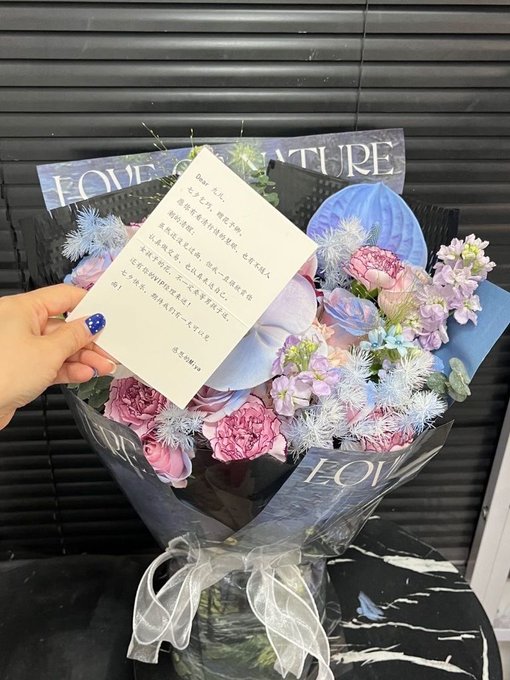 A hand holds a handwritten note in Chinese against a bouquet of pink and blue flowers wrapped in 'LOVE' and 'NATURE' themed paper.