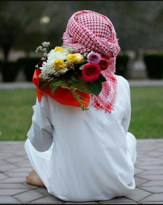 A photograph of a person sitting on paved ground, viewed from behind, wearing white robes and a red-and-white checkered keffiyeh headscarf, holding a bouquet of red, pink, yellow, and white flowers wrapped in red paper.