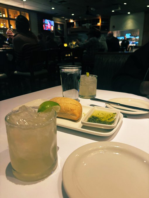 A margarita and bread with dipping oil sit on a white table at a dimly lit restaurant, with a TV and other diners visible in the background.

The image contrasts sharply with the post's reflection on financial hardship—showcasing an indulgent dining experience that serves as a visual symbol of recovery and progress after weathering a difficult financial period.