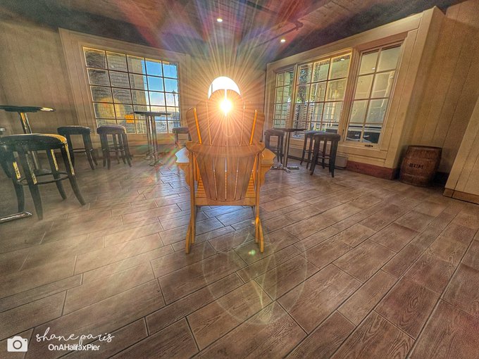 An interior view of a restaurant or bar with wooden floors, high-top tables and stools, large windows, and a wooden chair facing the sun, with watermark "shaneparis OnAHallmarkPier."