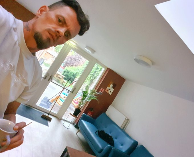 A man with a goatee wearing a white t-shirt stands in a living room, holding a small bowl, with a teal sofa and glass doors leading to a garden visible in the background.