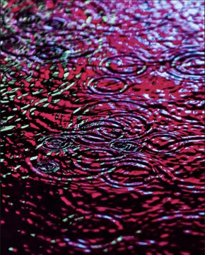 An abstract, swirling pattern of rippling textures in deep reds, magentas, and teal highlights, resembling a close-up of liquid or polished stone.