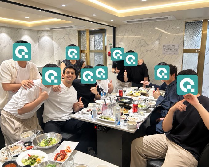 A group of people gather around a dining table filled with various dishes, posing for a photo with their faces obscured by teal logo icons.