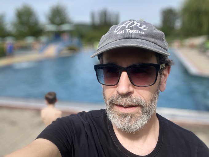 The image features a man wearing sunglasses, a black t-shirt, and a gray baseball cap with white text that reads "art on tozos". The man appears to be posing for a photo while standing near a pool or body of water. In the background, there are other people present, but they are not the main focus of the image.