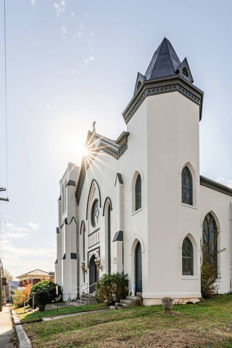 A white Gothic Revival church with a tall steeple and pointed arch windows stands bathed in golden sunlight, its grand yet weathered facade fitting the humorous tone of a quirky rental listing that playfully warns guests about fees for unauthorized exorcisms.