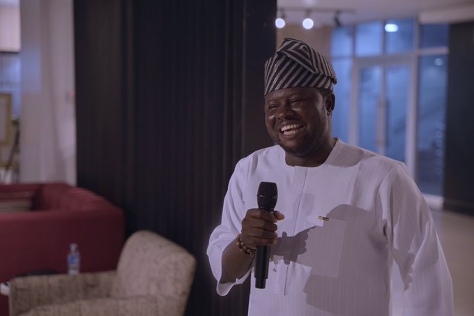 A man wearing traditional Nigerian attire, including a striped cap and white agbada, speaks into a microphone while smiling broadly in an indoor setting.