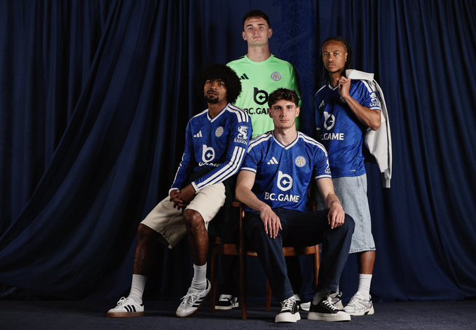 Four Leicester City football players posed in their 2025/26 kits (three in blue BC.GAME shirts and one goalkeeper in green) against a dark blue curtain backdrop.