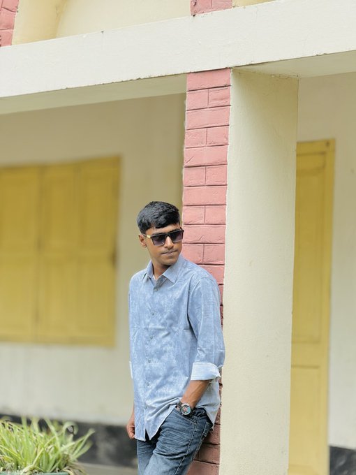 A young man wearing sunglasses, a light blue button-up shirt, and jeans leaning against a red brick pillar on the porch of a yellow building.