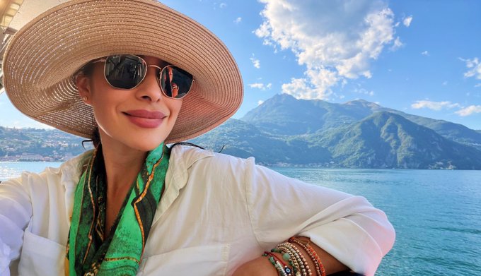 A woman wearing a large straw sun hat, sunglasses, and a white blouse with a green patterned scarf smiles at the camera, with a stunning lake and mountain landscape behind her. The scenic view complements the "Ciao Bella! Made it to paradise" caption, capturing the essence of an idyllic Italian getaway.