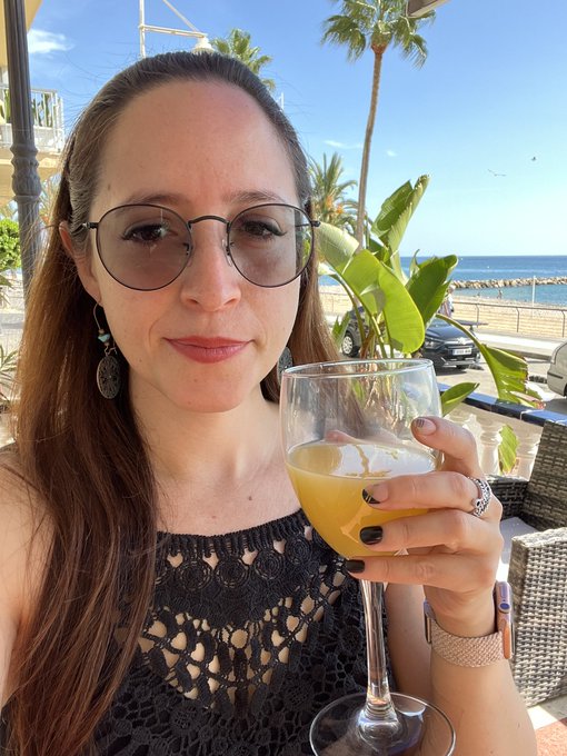 A selfie of a smiling woman with round glasses and a black crocheted top, holding a glass of yellow drink, with a sunny beach, palm trees, and the ocean visible behind her.