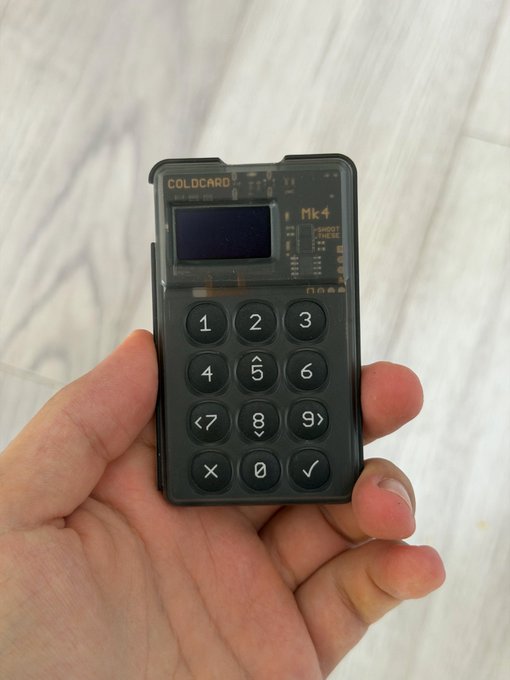 A hand holding a small black COLDCARD Mk4 hardware wallet device featuring a numeric keypad and display screen, used for secure cryptocurrency storage.