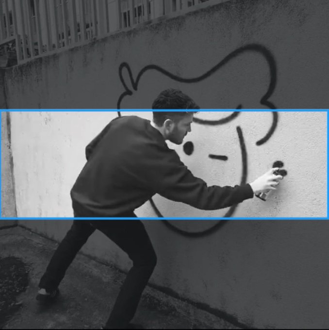 A black and white photo of a man spray-painting a graffitied character on a wall, with a blue rectangular frame highlighting the middle portion of the image.