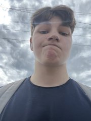 A close-up selfie of a young person with short hair wearing a dark shirt and backpack straps, standing outside against a cloudy sky.