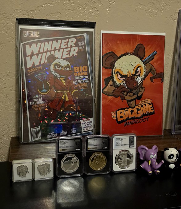 A collection of memorabilia is displayed, including a "Winner Winner Magazine" featuring a bandicoot character, a "Big Game Bandicoot" poster, several graded silver and gold coins in protective slabs, and small elephant, elephant, elephant, purple elephant, and panda figurines.
