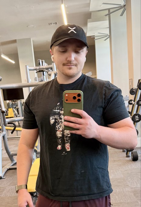 A man in a black cap and dark graphic t-shirt taking a mirror selfie with his phone in a gym setting.