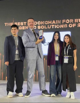 A photograph of four people posing on a stage at a blockchain event, with a backdrop reading 'THE BEST BLOCKCHAIN FOR [RE] GENERATIVE SOLUTIONS OF A...'.