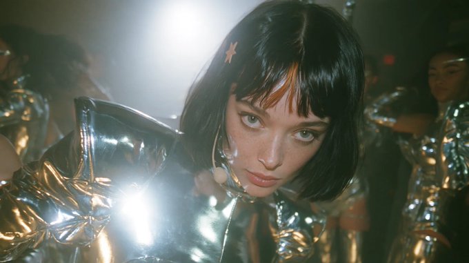 A young woman with a black bob haircut and orange bangs wears a shiny silver outfit in what appears to be a music video or performance scene.