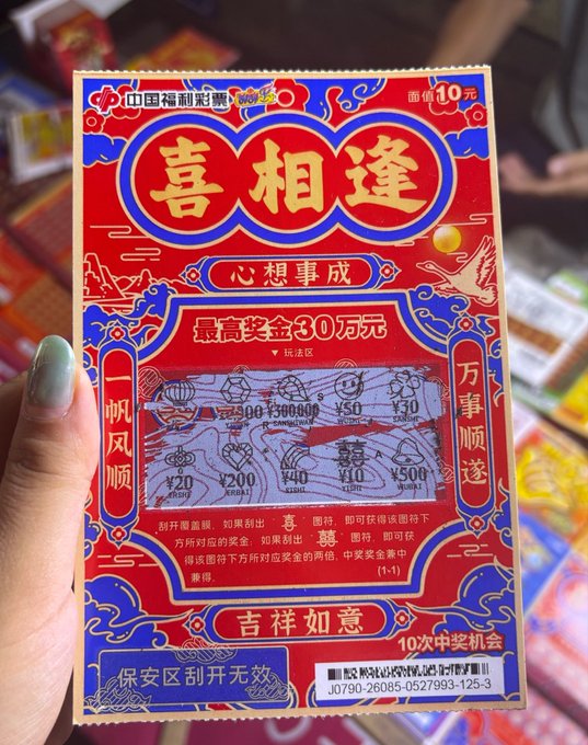 A hand holds a Chinese lottery scratch card (中国福利彩票) titled '喜相逢' (Joyful Reunion), featuring red and blue traditional designs and a scratch area with various prize amounts.