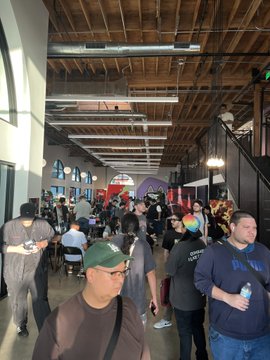 A crowded indoor venue with exposed wooden ceiling beams and industrial lighting, where many people are gathered, some wearing face masks, walking through what appears to be a convention or event space.