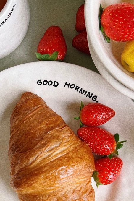 A white plate with the text 'GOOD MORNING.' holds a croissant and three fresh strawberries, accompanied by bowls of additional fruit.