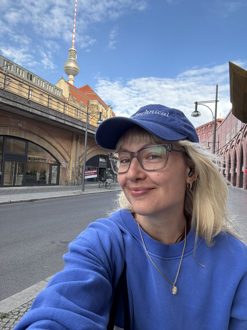 A woman with blonde hair wearing a blue sweatshirt and blue 'Astromical' cap takes a selfie in front of Berlin's Fernsehturm TV tower and a brick railway bridge.