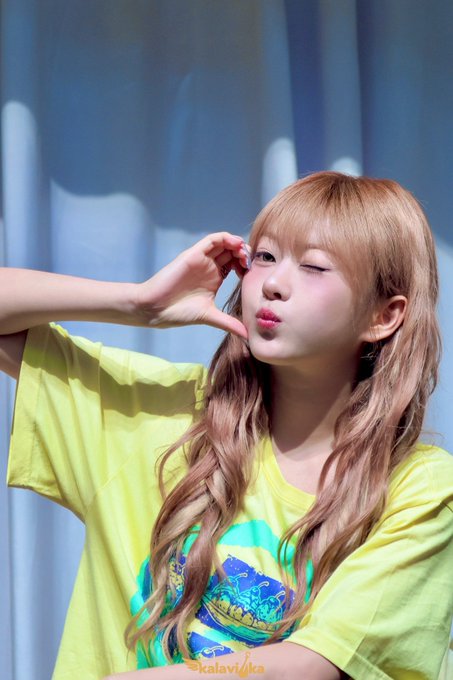 A portrait of a young woman with long wavy light brown hair wearing a yellow t-shirt, winking and making a heart gesture with her hands, credited to @kalaviyaka.