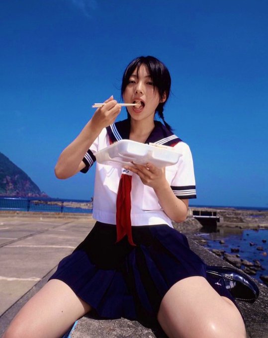 A young woman in a Japanese school uniform (sailor-style top with red tie and dark pleated skirt) sits outdoors by the sea, eating from a white takeout container with chopsticks.