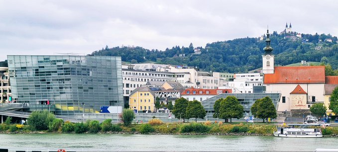 A cityscape view from across a river, featuring a modern glass building on the left, historic European architecture with a church spire on the right, and green hills in the background.