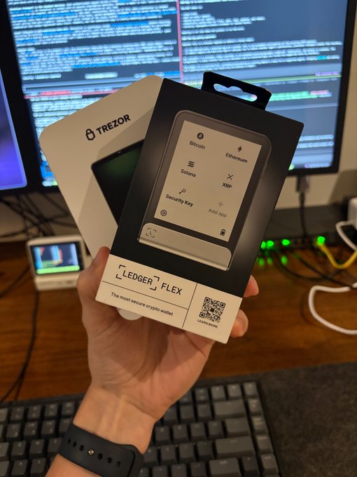 A hand holds a "LEDGER FLEX" hardware wallet box, billed as "The most secure crypto wallet," with a Trezor box visible behind it, set against a backdrop of code on a monitor—showing the hardware freshly unboxed for stress-testing on the anzen.sh MVP.