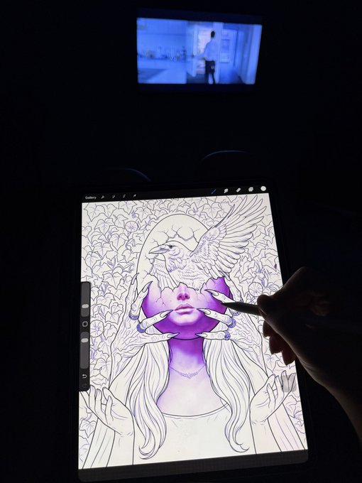 A hand uses a stylus on a tablet to digitally color a purple-toned illustration of a woman with a dove and clawed hands, while a small monitor in the background displays a blue-tinted video of a person.