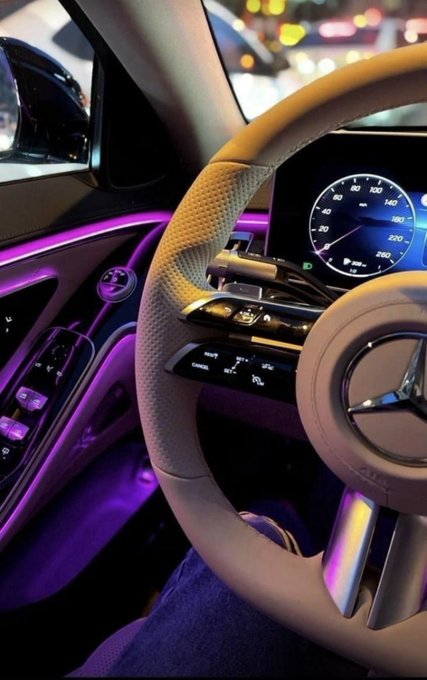 The interior of a Mercedes-Benz is shown, featuring a white steering wheel with the Mercedes logo, purple ambient lighting, and an illuminated digital speedometer displaying speeds up to 220.