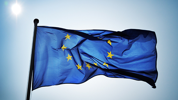 The European Union flag waving against a clear blue sky with the sun shining brightly in the background.