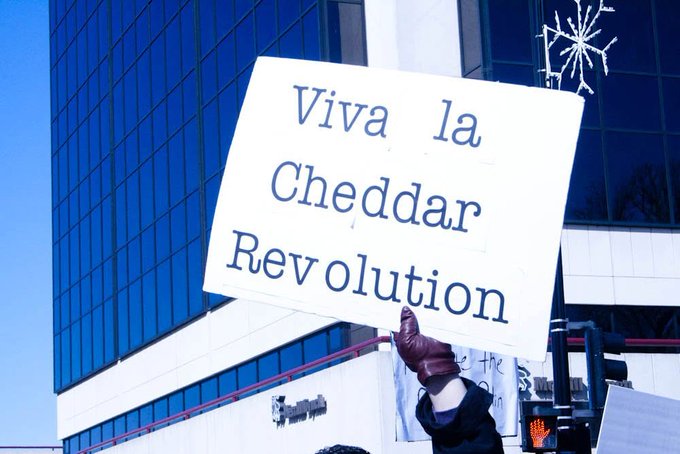 A person wearing a brown leather glove holds up a white protest sign reading 'Viva la Cheddar Revolution' in front of a blue glass office building.