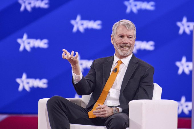 Michael Saylor, executive chairman of MicroStrategy, speaking at a CPAC event with a blue star-spangled backdrop, wearing a suit with an orange tie.