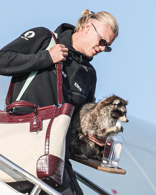 A woman with blonde hair wearing a black K-Way jacket and sunglasses, holding a raccoon that is drinking from a glass bottle of clear liquid on what appears to be a boat.