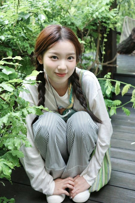 A young woman with braided hair crouching on a wooden deck surrounded by green foliage.