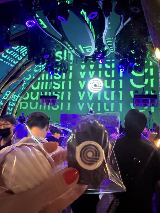A concert or event venue lit with neon green screens displaying abstract patterns, with a person in the foreground holding a plastic-wrapped cookie with an '@' symbol design.