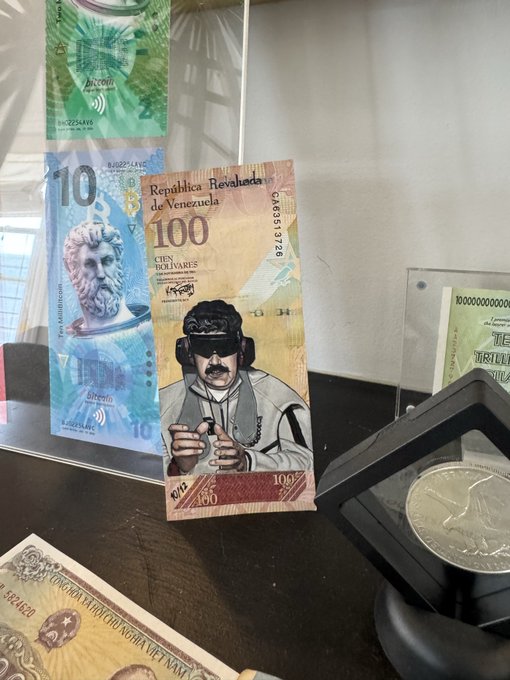 A display case showcases newly issued Venezuelan currency, including a 100 bolívar note featuring a portrait of Pablo de la Torriente Brau wearing headphones and sunglasses, alongside a 10 bolívar note depicting Ptolemy I. The image aligns with the caption by highlighting museum specimens of Venezuela's modernized banknotes, reportedly purchased with Bitcoin.