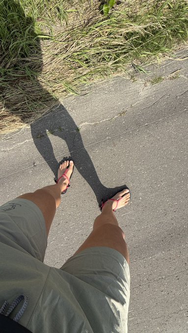 A first-person perspective looking down at a person's legs in gray shorts and pink flip-flops, casting a long shadow on the concrete path beside grassy terrain.