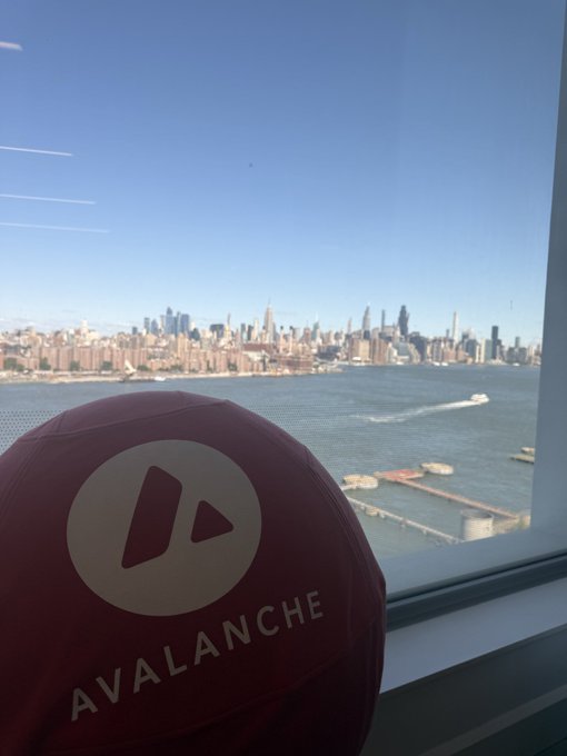 A maroon baseball cap with the 'AVALANCHE' logo rests in the foreground, with a city skyline and body of water visible through a window in the background.