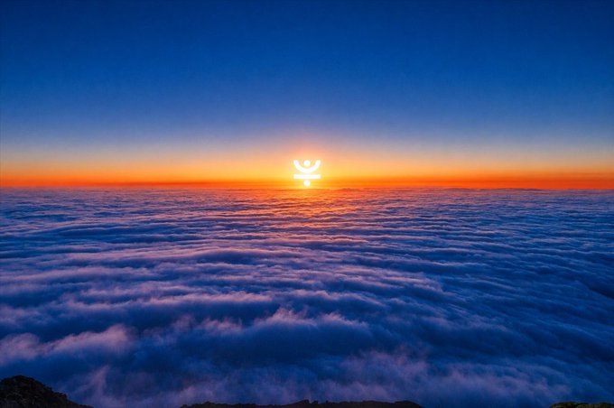A scenic view shows a sunset above a sea of clouds with blue sky, featuring a glowing sun symbol in the center.