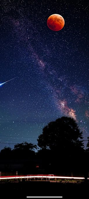 A vertical photograph of a brilliant red blood moon hanging in a starry night sky above the Milky Way, with silhouetted trees and light trails from passing cars in the foreground.