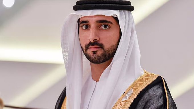 A man wearing traditional Emirati attire, including a white ghutra with a black agal and a dark bisht with gold trim, looking directly at the camera.