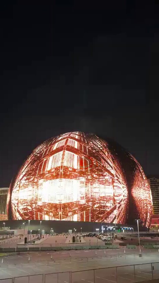 The image features a large, illuminated ball structure that appears to be a dome or a building with a unique design. The structure is lit up at night, making it stand out against the dark sky. The photo has been captioned by @DavidAriew, who wrote about the image on his social media account. The post also includes a link to the original source of the image, which is a website called Render Network.