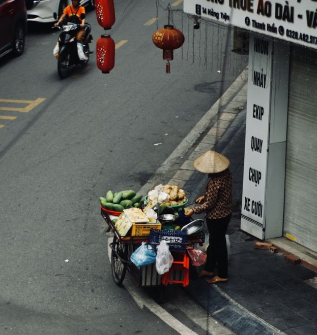 A street vendor wearing a traditional conical hat pushing a cart of produce, including mangoes, down a street in Vietnam near a shop with red lanterns.