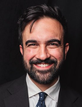 A smiling, bearded man in a dark suit and polka-dot tie poses against a black background, accompanying news about Mamdani's comments on his political disagreements with Trump while sharing love for New York City, with a tongue-in-cheek 31% prediction for Time Person of the Year.