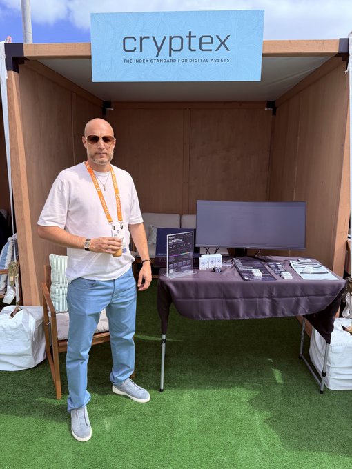 A bald man wearing sunglasses, a white T-shirt, light blue pants, and gray shoes stands at the cryptex booth ("THE INDEX STANDARD FOR DIGITAL ASSETS") with a computer monitor and branded materials on display at an outdoor conference.