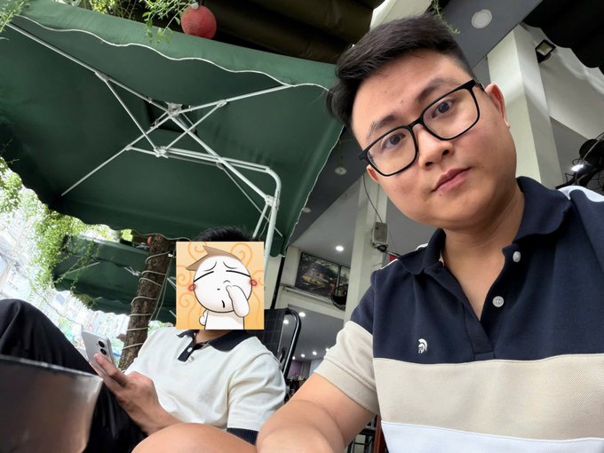 A selfie of an Asian man in glasses wearing a navy and white polo shirt, sitting under a green umbrella at an outdoor café with another person whose face is covered by a cartoon sticker.